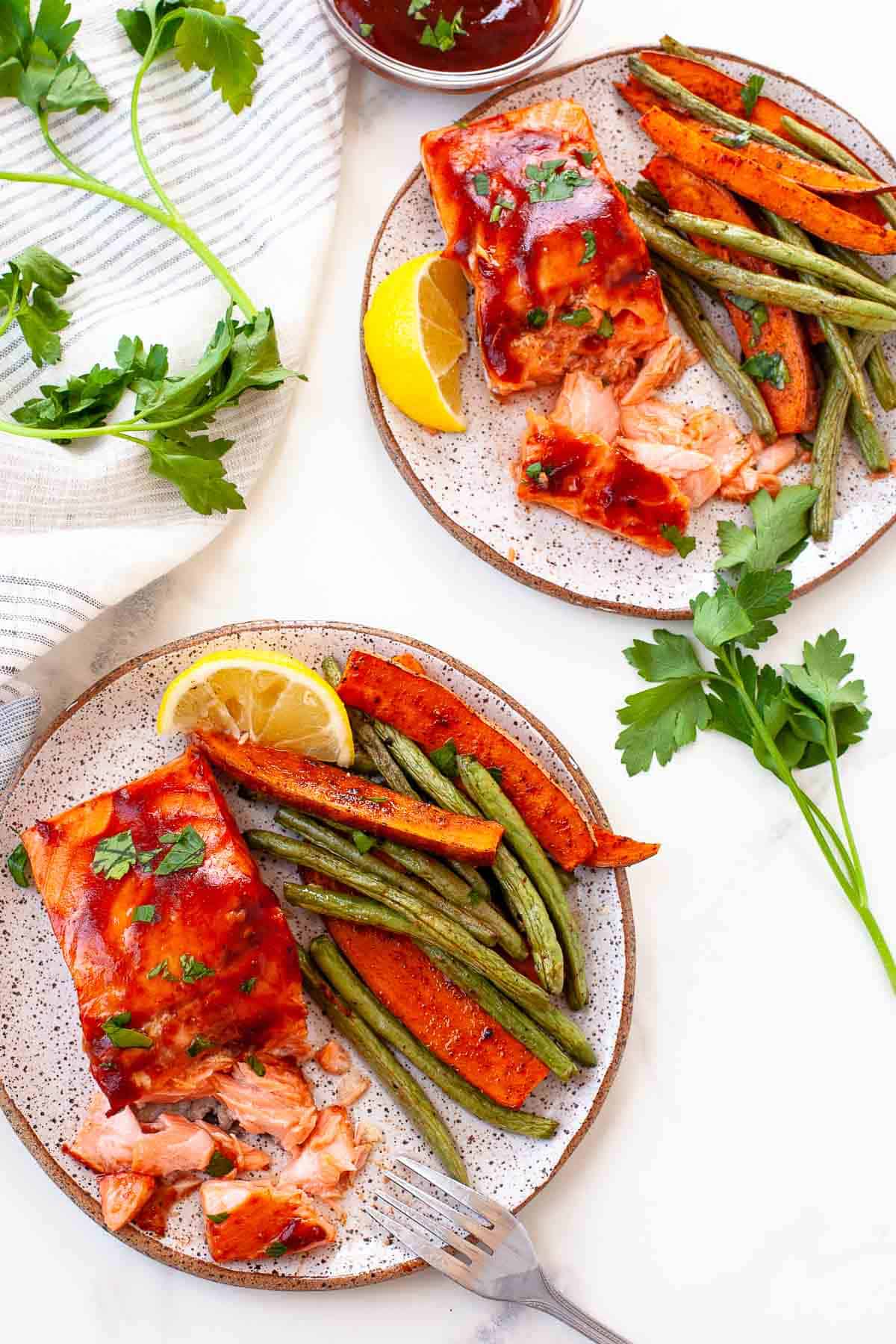 two plates bbq salmon. flaked. green beans. sweet potato. lemon slice. sprigs of cilantro. striped napkin. extra bbq sauce. 