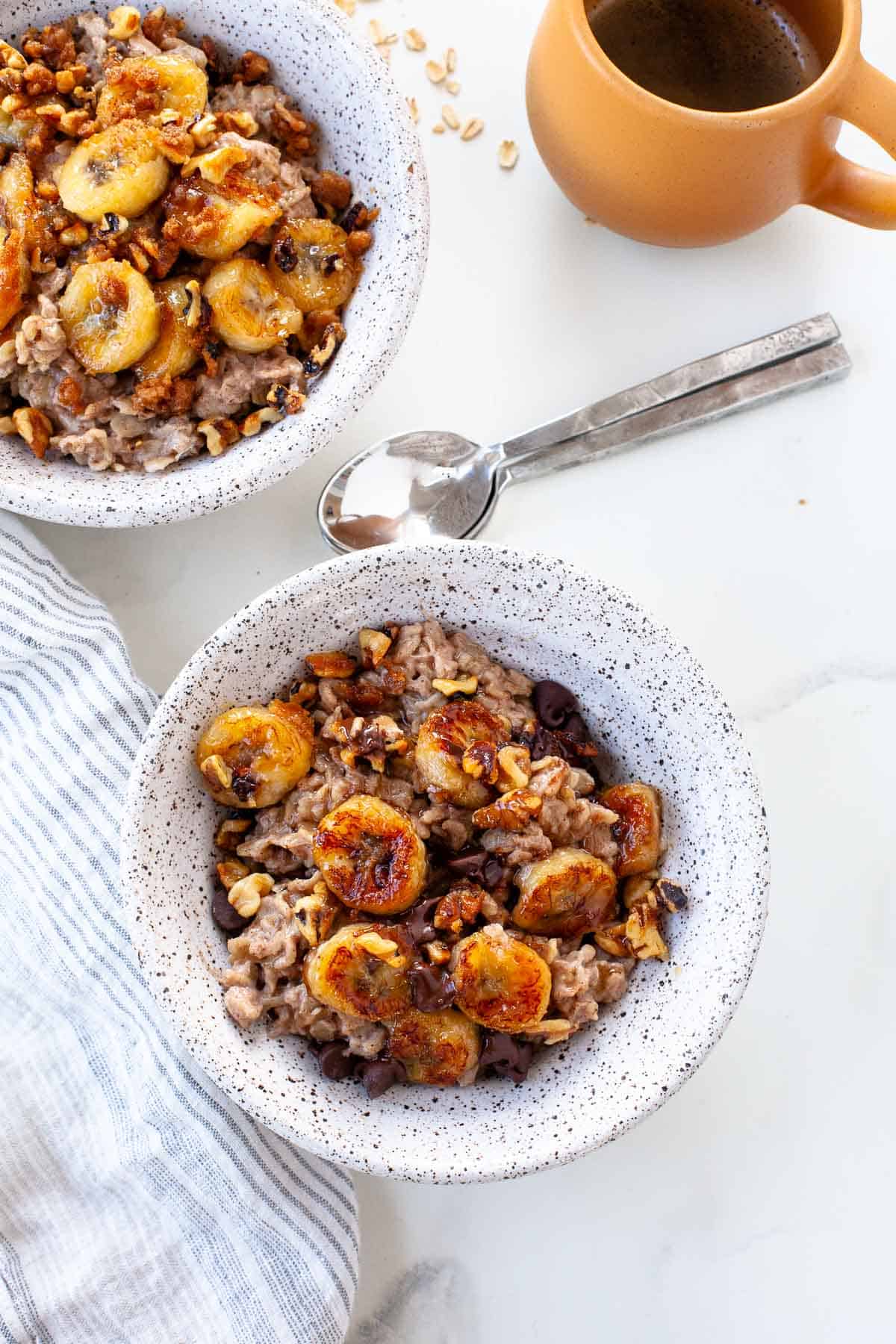 two banana bread oatmeal in white speckled ceramic bowls over white marble background next to orange coffee mug and navy striped napkin and silver spoons