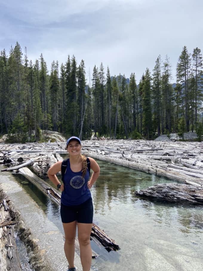 woman with hat in shorts and tank top hiking with stream, logs, and trees in background 