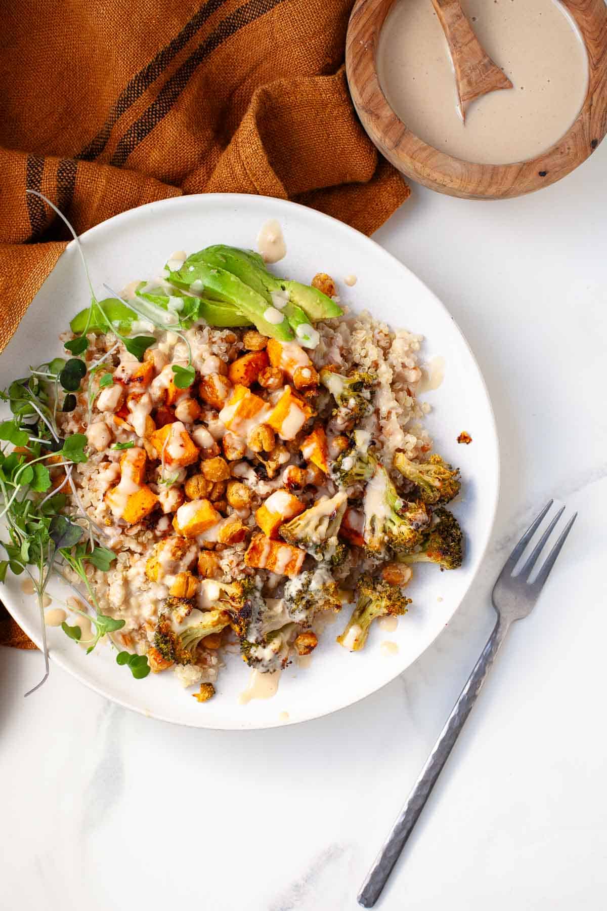 Ancient grains bowl on white marble surface beside orange kitchen towel and small wooden bowl of creamy tahini sauce.