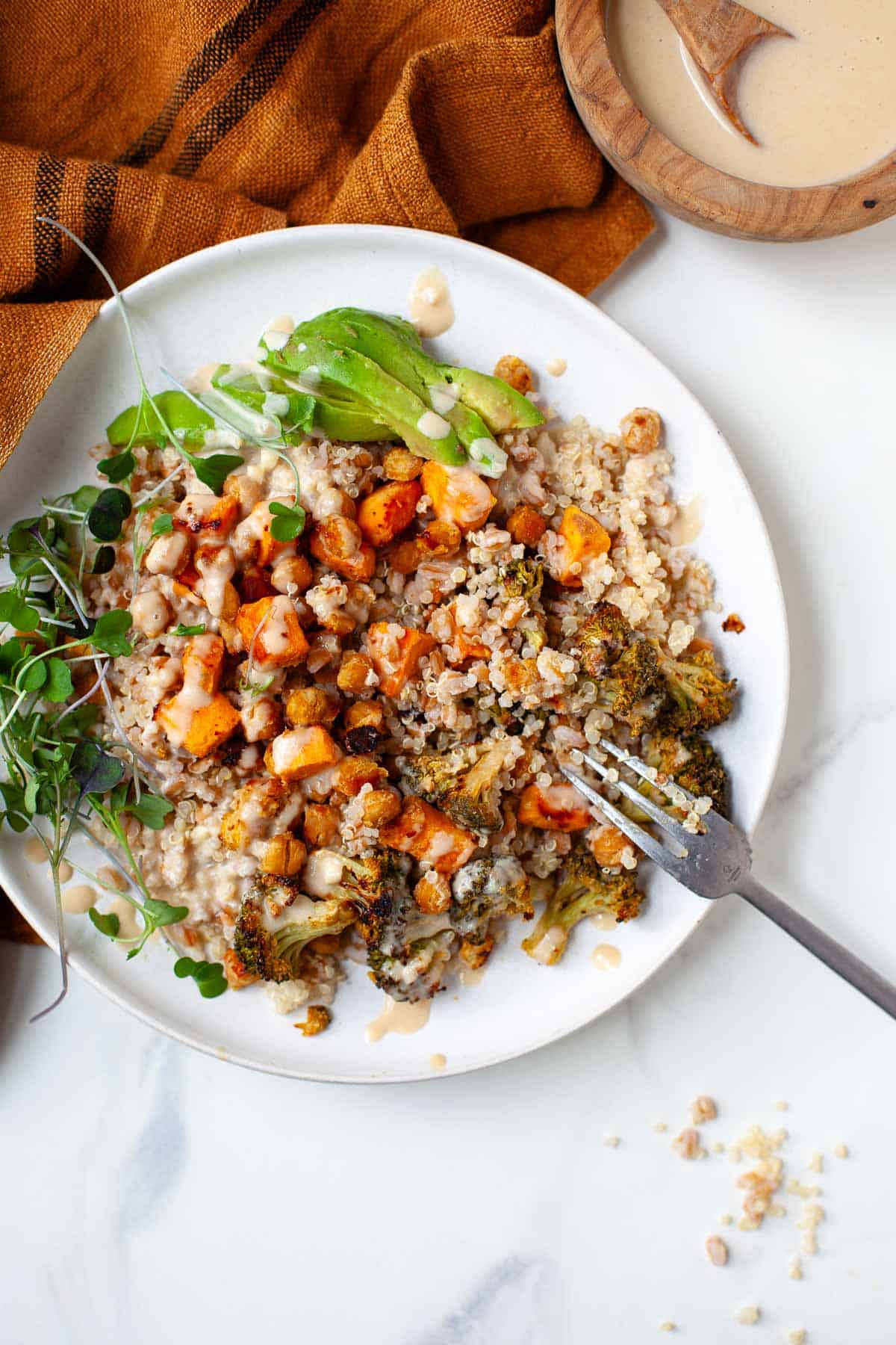 Ancient grains bowl in white bowl garnished with microgreens next to orange kitchen towel and wood bowl with tahini sauce