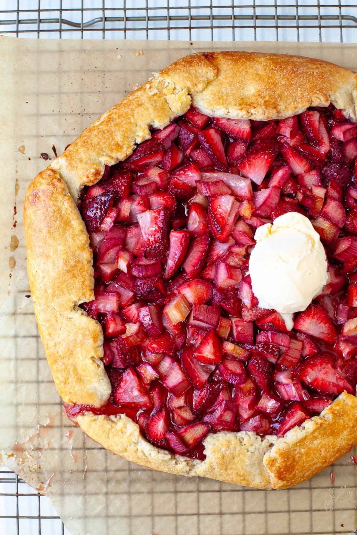 strawberry rhubarb galette topped with a scoop of vanilla ice cream on a parchment paper lined wire rack.