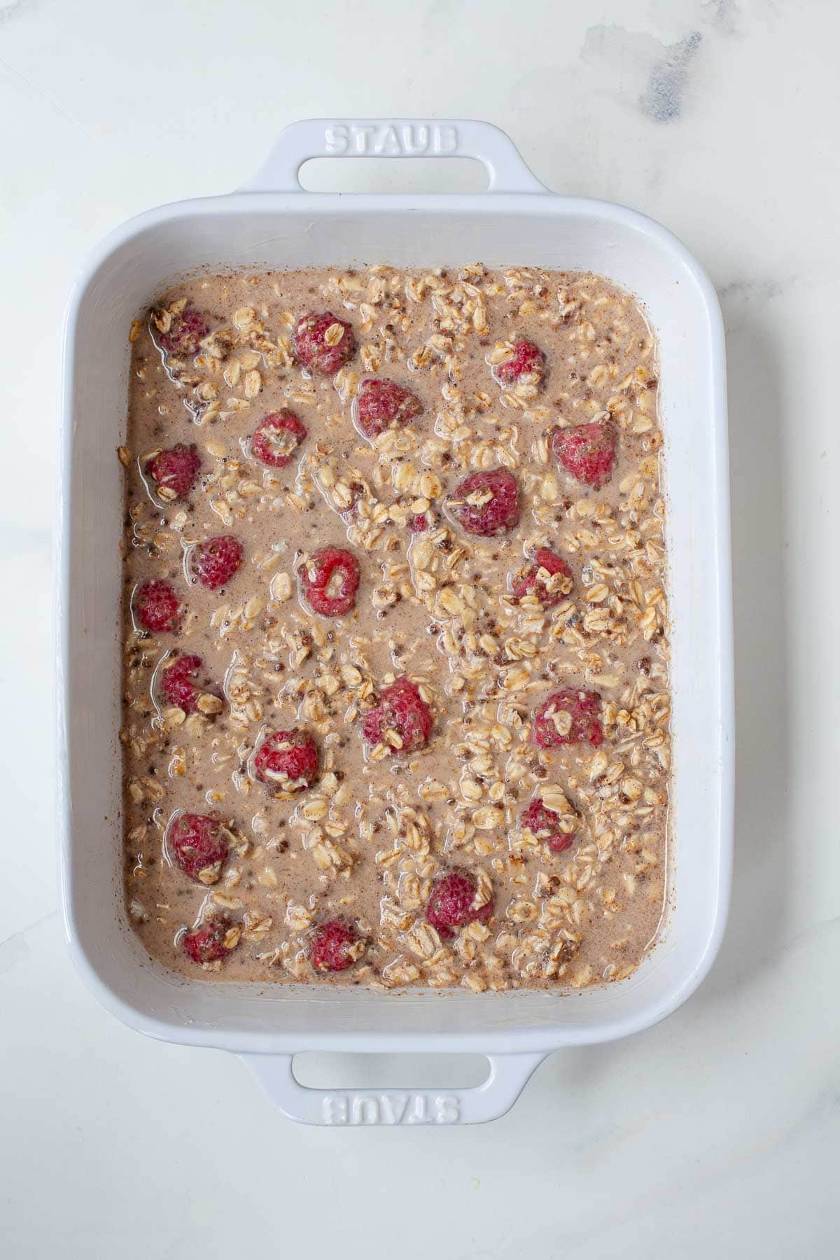 Prepped vegan oatmeal mixture with raspberries in white ceramic baking dish on marble counter. 